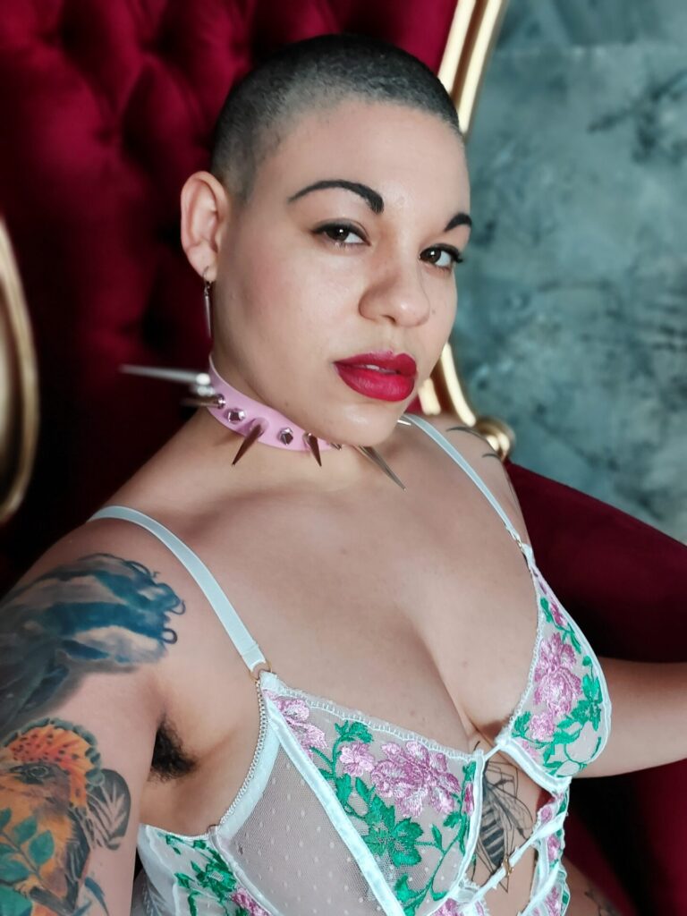 Oran Julius, a bald Domme, is sitting in a throne wearing mesh lingerie and a pink spiked collar.