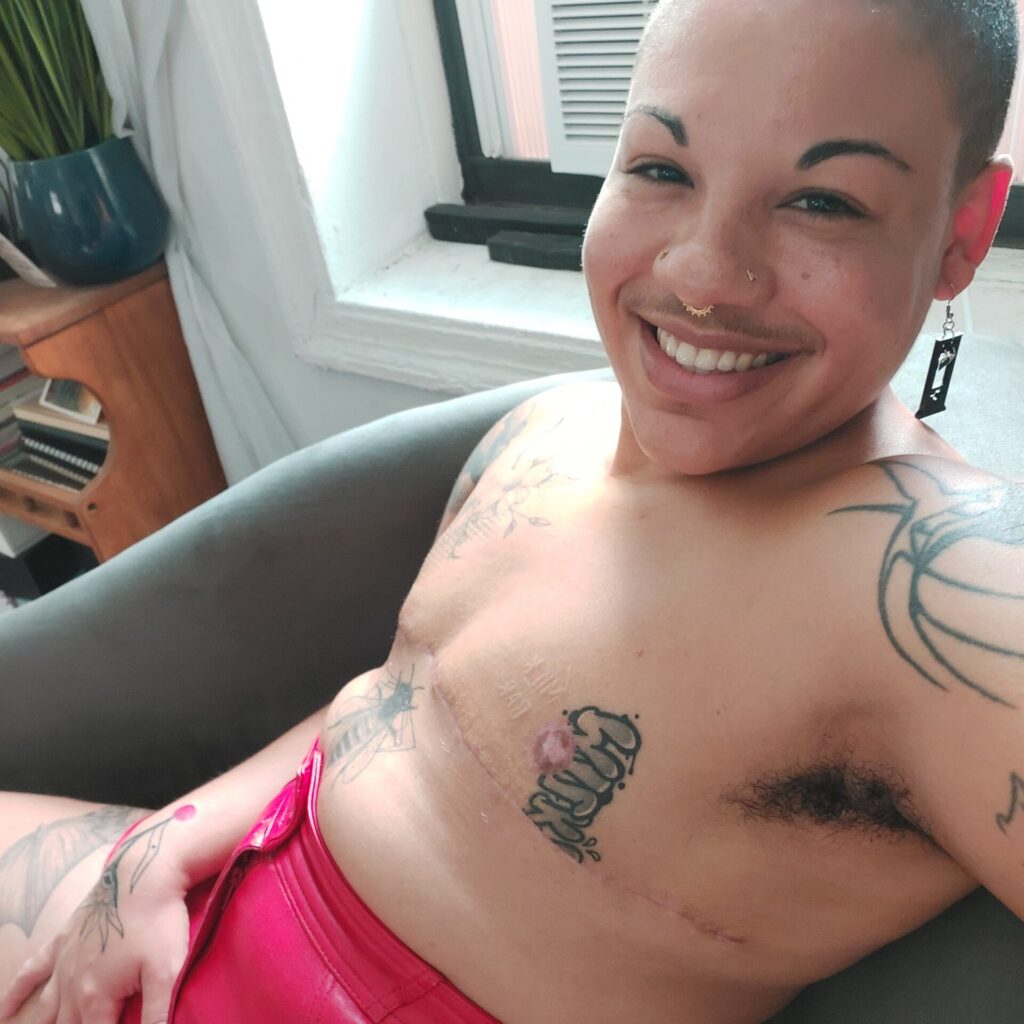 Oran Julius a FTM Domme with a shaved head is lounging in red leather and shirtless, displaying their top surgery scars and tattoos.