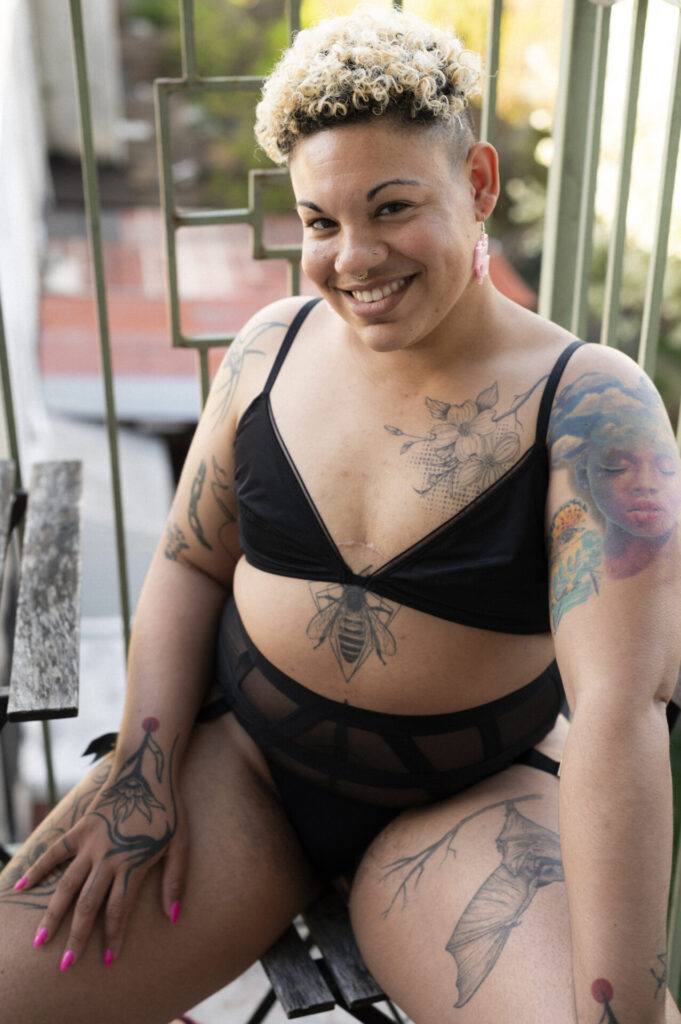 Oran Julius, nonbinary femme Dominant posing with a gorgeous smile in the summer sun